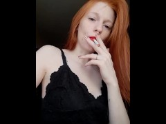 A girl with long red hair in a black dress and red lipstick smokes a cigarette with a brown filter