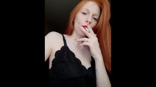 A girl with long red hair in a black dress and red lipstick smokes a cigarette with a brown filter